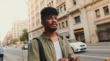 Young smiling man with beard dressed in an olive color shirt uses phone map app on the old city background