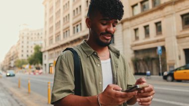 Young smiling man with beard dressed in an olive color shirt uses phone map app on the old city background