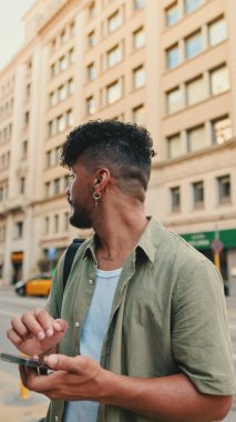 Young smiling man with beard dressed in an olive color shirt uses phone map app on the old city background