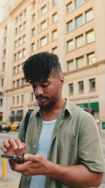 Young smiling man with beard dressed in an olive color shirt uses phone map app on the old city background