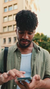 Young smiling man with beard dressed in an olive color shirt uses phone map app on the old city background