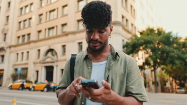 Young smiling man with beard dressed in an olive color shirt uses phone map app on the old city background