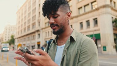 Young smiling man with beard dressed in an olive color shirt uses phone map app on the old city background