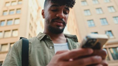 Young smiling man with beard dressed in an olive color shirt uses phone map app on the old city background