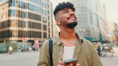 Young smiling man with beard dressed in an olive color shirt uses phone map app on the old city background