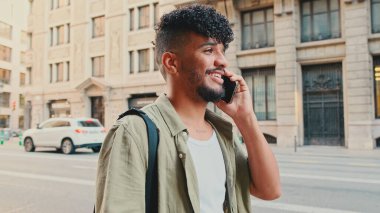 Young happy man with beard dressed in an olive-colored shirt is talking on cellphone standing on the old city background