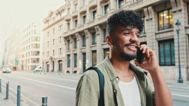 Young happy man with beard dressed in an olive-colored shirt is talking on cellphone standing on the old city background