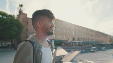 Young man dressed in an olive-colored shirt is standing in the port looking at the yachts