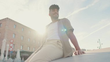 Zeytin rengi gömlek giyen genç adam limandaki rıhtımda oturuyor. 