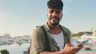 Young man dressed in an olive-colored shirt is sitting on the waterfront in the port watching videos and photos on his cellphone.