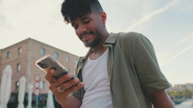 Young man dressed in an olive-colored shirt is sitting on the waterfront in the port watching videos and photos on his cellphone.