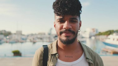 Zeytin rengi gömlek giymiş sakallı mutlu genç adam yatların yanında kameraya bakıyor.