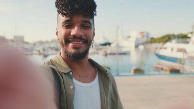 Sakallı, zeytin rengi tişört giyen genç adam yat limanının yanında duruyor, gülümsüyor ve selfie çekiyor.
