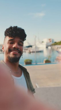 Sakallı, zeytin rengi tişört giyen genç adam yat limanının yanında duruyor, gülümsüyor ve selfie çekiyor.