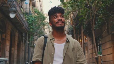 Close up, young smiling man dressed in an olive-colored shirt walks along the narrow street of the old city and looks around