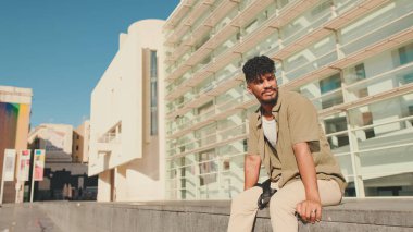 Young male student dressed in an olive colored shirt sits outside next to university