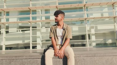 Young male student dressed in an olive colored shirt sits outside next to university