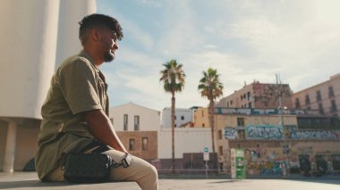 Young male student dressed in an olive-colored shirt sits outside next to the university, listening to music on headphones