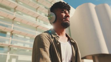 Young male student dressed in an olive-colored shirt sits outside next to the university, listening to music on headphones