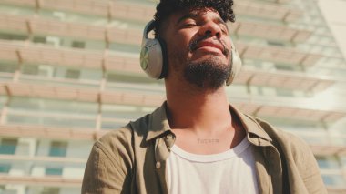 Young male student dressed in an olive-colored shirt sits outside next to the university, listening to music on headphones