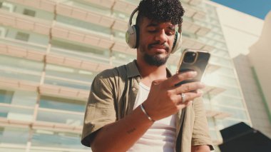 Close up, young male student dressed in an olive color shirt sits outside next to the university, listens to music on headphones, selects tracks on his phone