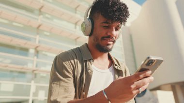 Close up, young male student dressed in an olive color shirt sits outside next to the university, listens to music on headphones, selects tracks on his phone