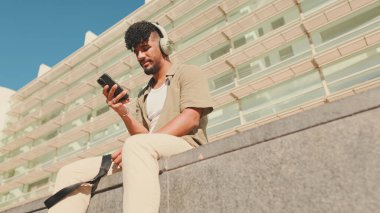 Close up, young male student dressed in an olive color shirt sits outside next to the university, listens to music on headphones, selects tracks on his phone
