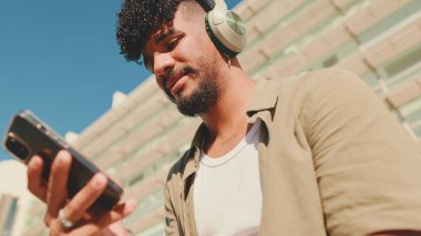 Close up, young male student dressed in an olive color shirt sits outside next to the university, listens to music on headphones, selects tracks on his phone