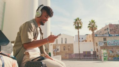 Close up, young male student dressed in an olive color shirt sits outside next to the university, listens to music on headphones, selects tracks on his phone