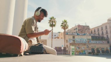 Close up, young male student dressed in an olive color shirt sits outside next to the university, listens to music on headphones, selects tracks on his phone
