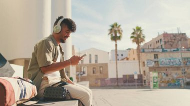 Close up, young male student dressed in an olive color shirt sits outside next to the university, listens to music on headphones, selects tracks on his phone
