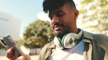 Young man with beard wearing an olive-colored shirt with headphones sits on bench and uses cellphone