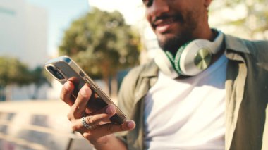 Young man with beard wearing an olive-colored shirt with headphones sits on bench and uses cellphone