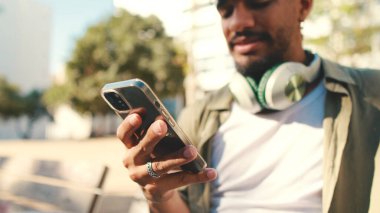 Young man with beard wearing an olive-colored shirt with headphones sits on bench and uses cellphone
