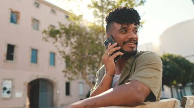 Young man with beard wearing an olive colored shirt with headphones talking on the phone while sitting on bench on modern buildings background