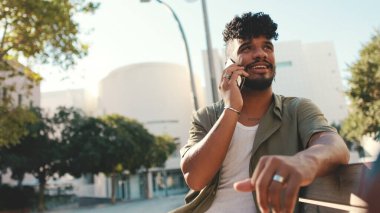 Young man with beard wearing an olive colored shirt with headphones talking on the phone while sitting on bench on modern buildings background
