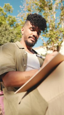Young male student wearing an olive colored shirt takes notes in notepad while sitting on bench outside the university