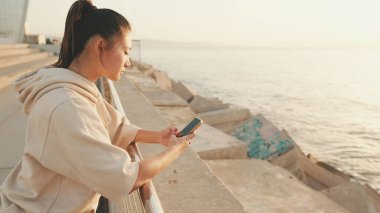 Smiling asian girl in sportswear uses phone while standing on waterfront on modern buildings background in morning light