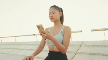 Close-up of smiling asian girl in sports top using cellphone while standing on waterfront on modern buildings background in morning light