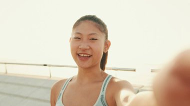 Smiling asian girl in sports top makes selfie while standing on the embankment in the morning light