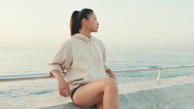 Asian girl wearing sportswear sits on the waterfront, enjoys the fresh air at morning time