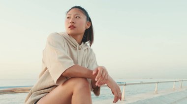 Asian girl wears sportswear sits on the promenade at morning time, on the background of the sea
