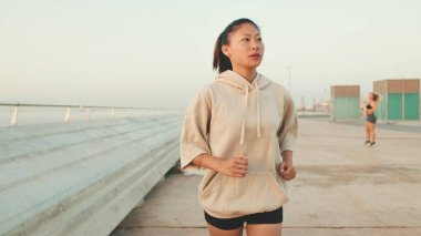 Asian young athletic woman running along the promenade. Sportswoman in sportswear exercising outdoors at morning time