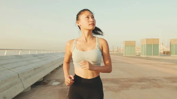 Asian girl in sports top runs along the promenade at morning time