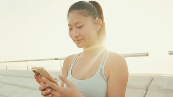 Close-up of smiling asian girl in sports top using cellphone while standing on waterfront on modern buildings background in morning light