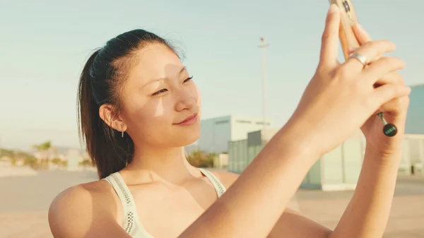 Asian girl in sports top makes selfie while standing on the embankment on modern buildings background in the morning light