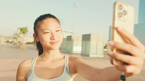 Asian girl in sports top makes selfie while standing on the embankment on modern buildings background in the morning light