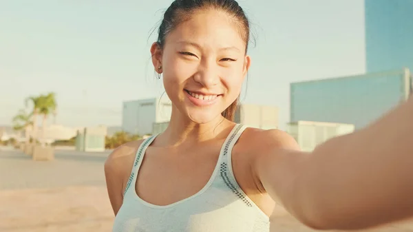 Smiling asian girl in sports top makes selfie while standing on the embankment in the morning light