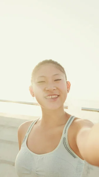 Smiling asian girl in sports top makes selfie while standing on the embankment in the morning light