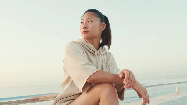 Asian girl wears sportswear sits on the promenade at morning time, on the background of the sea